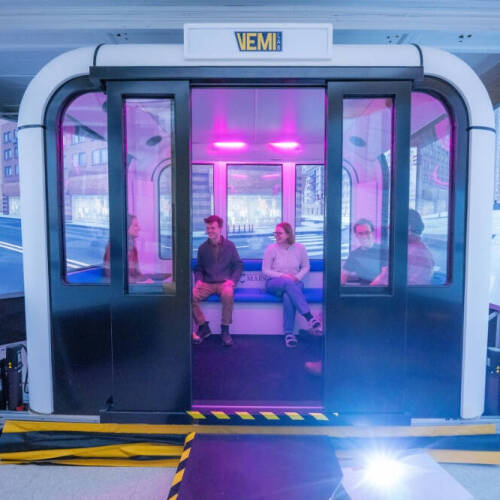 Two people sit inside a modern, enclosed vehicle simulator with sliding glass doors and purple lighting. The simulator is surrounded by large screens displaying a virtual cityscape, creating the illusion of being in an urban environment.
